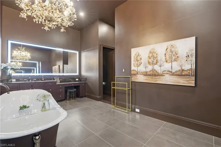 Freestanding soaking tub, dual vanity with undermount sinks, illuminated mirror, dark cabinetry, and tile flooring