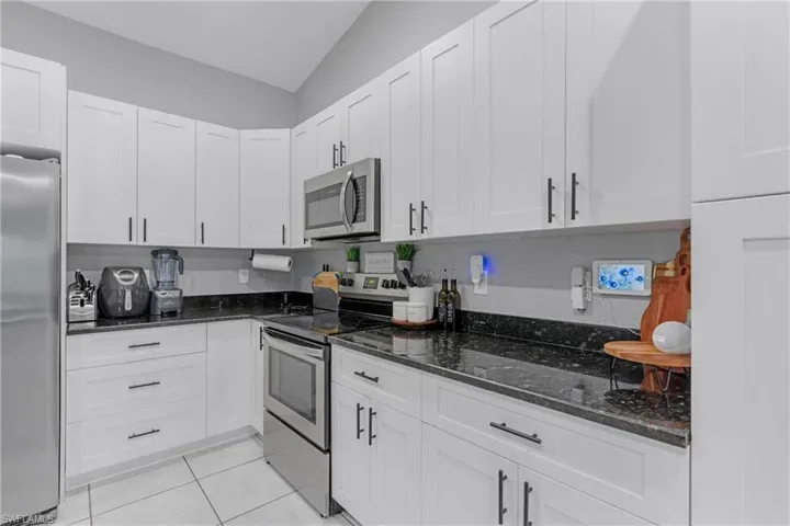 Kitchen featuring vaulted ceiling, dark stone counters, stainless steel appliances, white cabinets, and light tile floors