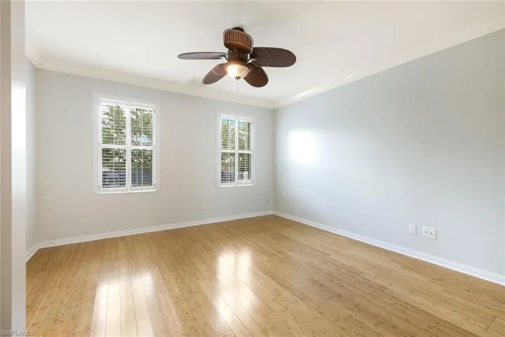 Spacious interior room featuring wood-finish flooring, two windows with blinds, a ceiling fan with integrated lighting, crown molding, and white baseboards