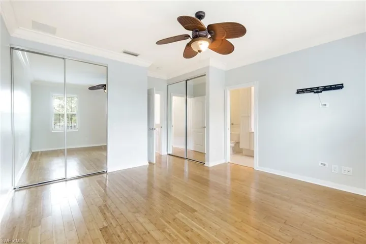 Spacious room featuring wood-finish flooring, mirrored closet doors, a ceiling fan with light, crown molding, and an attached bath