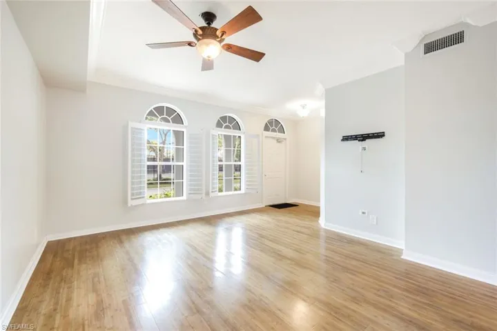 Spacious living area featuring wood-finish flooring, three arched windows with plantation shutters, a ceiling fan with integrated lighting, and crown molding