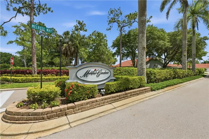 Melrose Gardens entrance featuring a curvilinear sign, stone-finish retaining wall, and manicured landscaping with mature trees