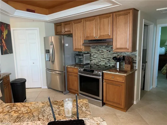 Kitchen featuring brown cabinetry, stainless steel appliances, light stone counters, light tile patterned flooring, and under cabinet range hood