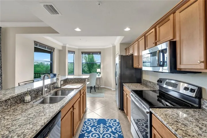 Kitchen featuring stainless steel appliances, crown molding, light stone counters, light tile patterned floors, and brown cabinetry