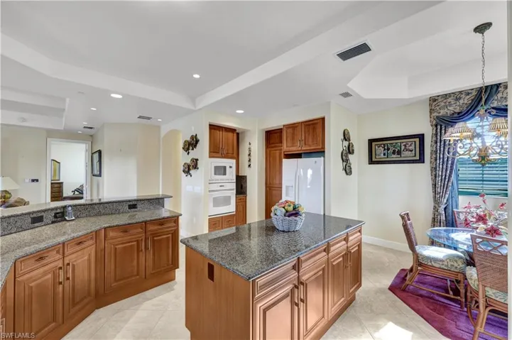 Kitchen with a raised ceiling, brown cabinets, dark stone counters, white appliances, and recessed lighting