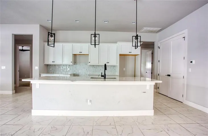 Spacious kitchen featuring an expansive island with a light-toned countertop, white cabinetry with dark hardware, a decorative tile backsplash, and three pendant light fixtures