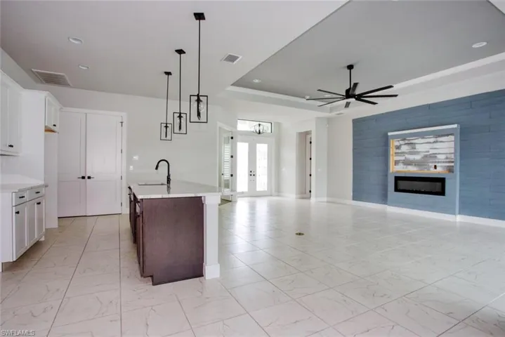 Spacious interior featuring large format tile flooring, a kitchen island with sink, white shaker cabinetry, a ceiling fan, and an accent wall with an inset electric fireplace