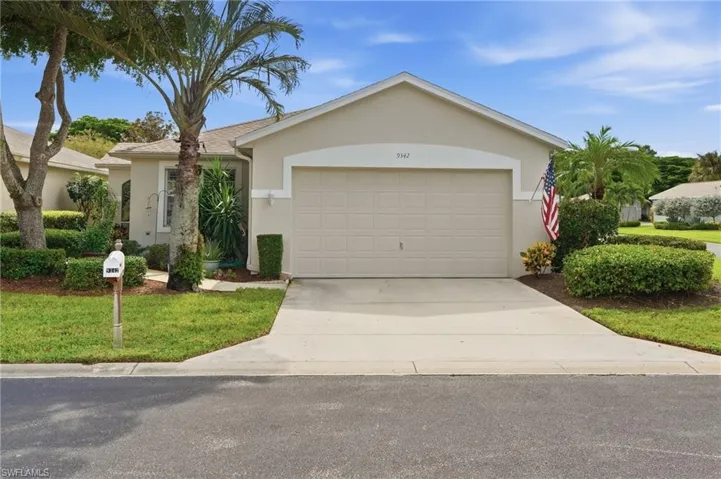 Stucco exterior with a pitched roof, attached garage, concrete driveway, and landscaped front yard with mature trees and shrubbery