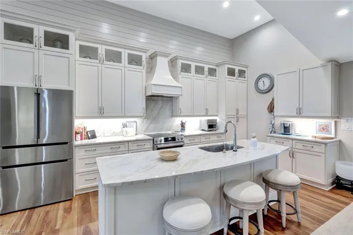 Kitchen with stainless steel appliances, custom range hood, light stone countertops, light wood-style flooring, and an island with sink