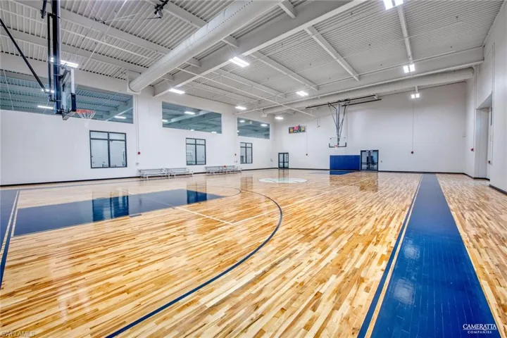 Indoor gymnasium featuring a wood-finish floor with court markings, high ceilings, multiple basketball hoops, recessed lighting, and upper-level windows