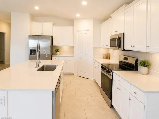 Modern kitchen featuring a large island with an undermount sink, stainless steel appliances, white shaker cabinetry, light-toned countertops, and a subtle geometric tile backsplash