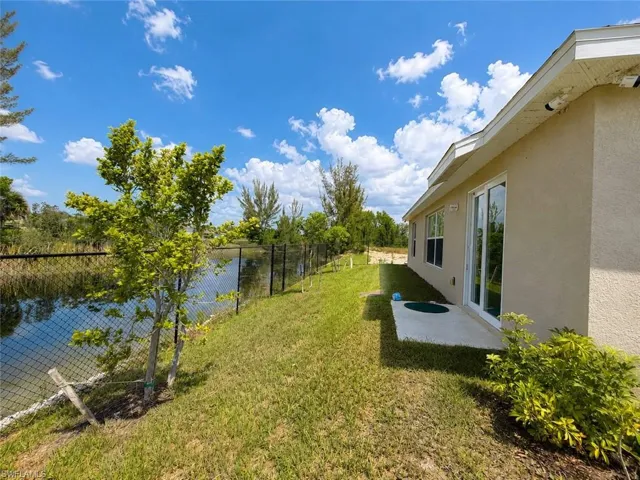 Expansive backyard featuring a water body, chain-link fencing, a concrete patio, mature trees, and a stucco exterior