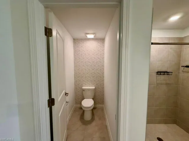 Full bathroom featuring a commode alcove with patterned wall covering, a built-in shower with tile surround, and neutral tile flooring