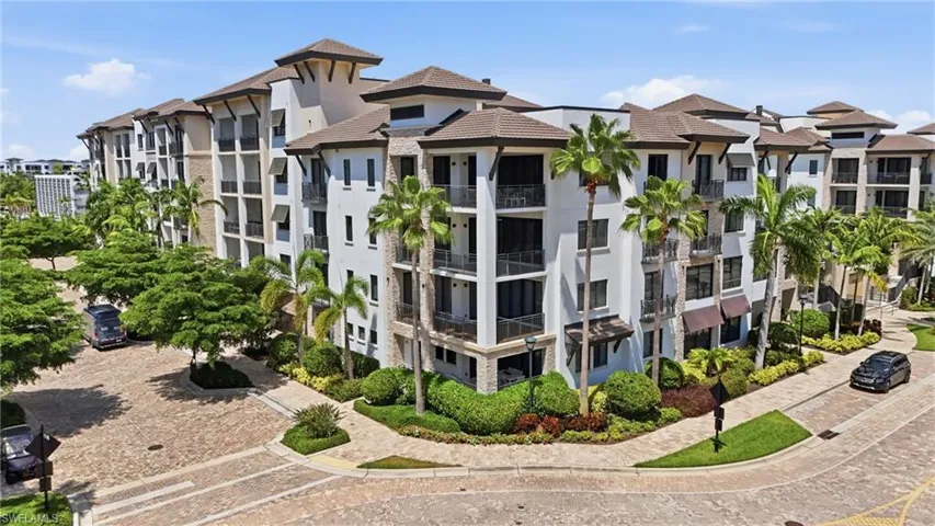 Multi-story residential building with private balconies and awnings, featuring a paver brick driveway, lush landscaping, and mature palm trees