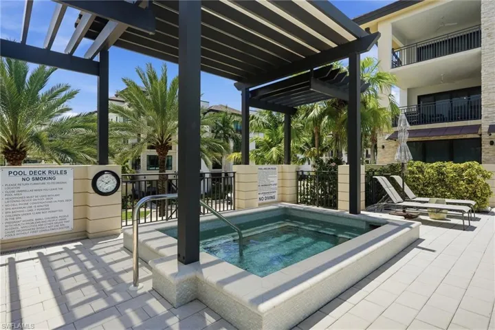 Spa with perimeter tiling under a dark pergola, surrounded by light-colored paver patio and tropical landscaping
