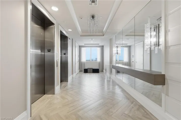 Stainless steel elevators, herringbone-patterned flooring, mirrored wall with floating ledge, contemporary chandelier, and recessed lighting