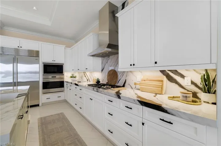 Kitchen featuring built in appliances, white cabinetry, light stone countertops, wall chimney range hood, and decorative backsplash