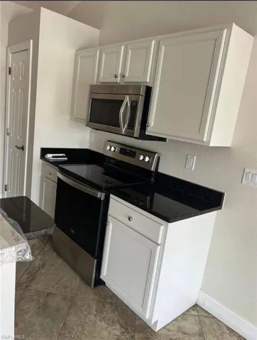 Kitchen featuring stainless steel appliances, white cabinets, a textured wall, light tile patterned floors, and dark stone countertops