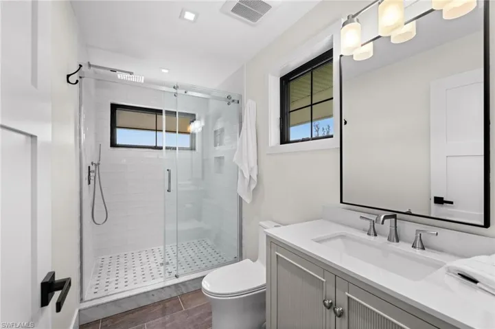 Vanity with integrated sink and chrome fixtures, framed mirror with task lighting, glass-enclosed shower with white subway tile surround, patterned shower floor, and a window with black trim
