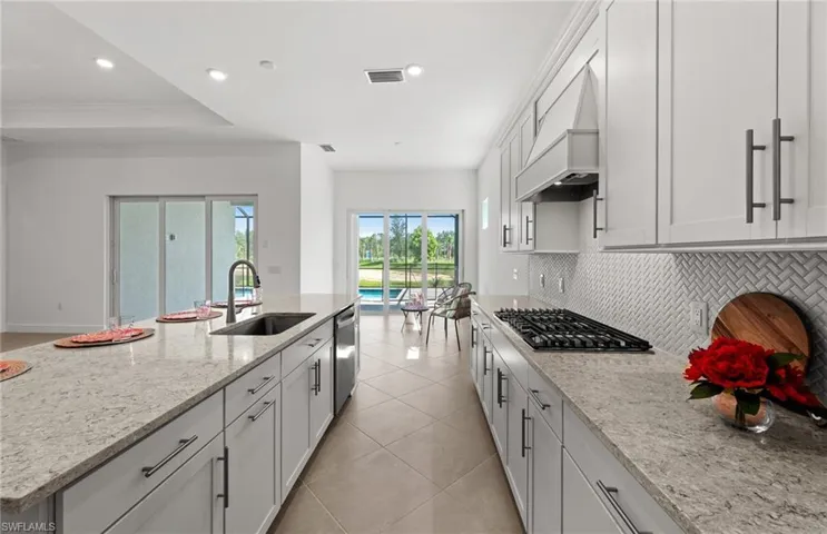 Kitchen with light tile flooring, a center island with sink, tasteful backsplash, custom exhaust hood, and appliances with stainless steel finishes