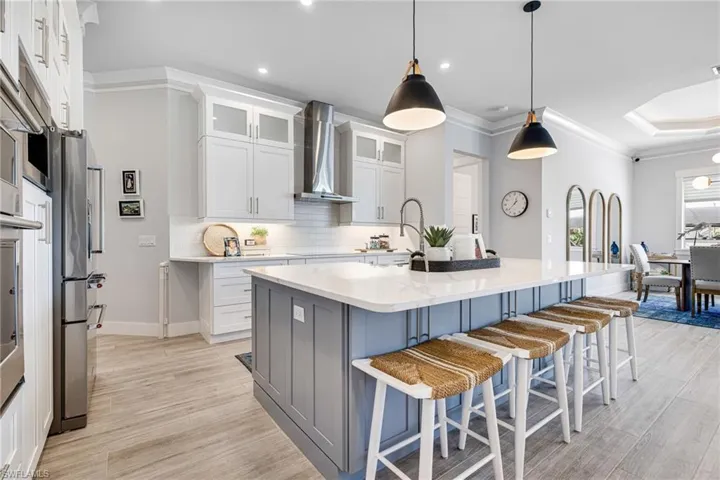 Kitchen with wall chimney exhaust hood, glass insert cabinets, kitchen island with sink, light cabinetry, quartz counters, and pendant lighting
