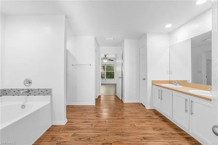 Spacious bathroom featuring wood-finish flooring, a built-in tub with tile surround, a dual vanity with white cabinetry, and a large wall-mounted mirror