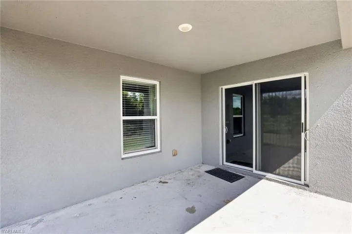 Covered outdoor patio with a concrete surface, featuring a large window with blinds and a sliding glass door