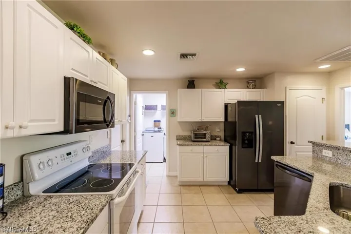 Kitchen with electric stove, fridge with ice dispenser, black microwave, white cabinets, and light stone countertops