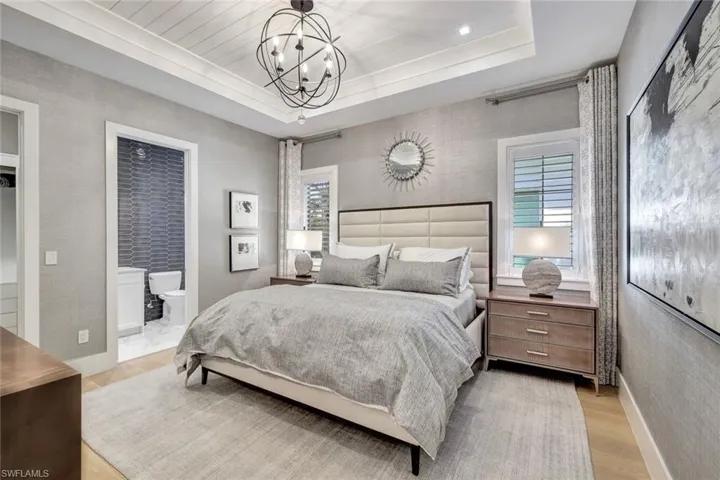 Bedroom with light wood-type flooring, a tray ceiling, ensuite bath, and an inviting chandelier