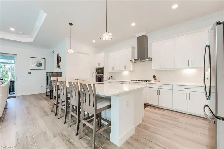 Kitchen with wall chimney exhaust hood, stainless steel appliances, white cabinetry, hanging light fixtures, and an island with sink