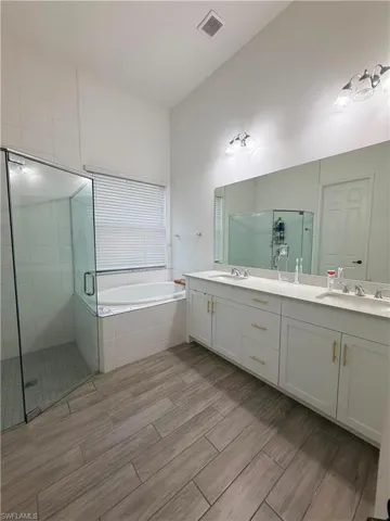 Bathroom featuring a dual vanity with white cabinetry and gold-tone hardware, a large frameless mirror, a glass-enclosed shower, a corner soaking tub, and wood-finish tile flooring