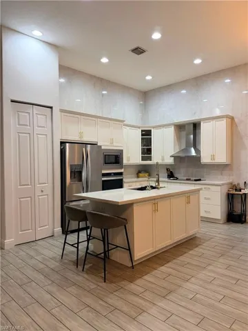 Modern kitchen featuring high ceilings with recessed lighting, an expansive central island with countertop seating, stainless steel appliances, white cabinetry with gold-tone hardware, and wood-finish tile flooring