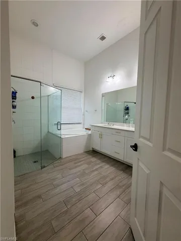 Bathroom featuring a glass-enclosed shower, corner soaking tub, double vanity with white cabinetry, and wood-finish tile flooring