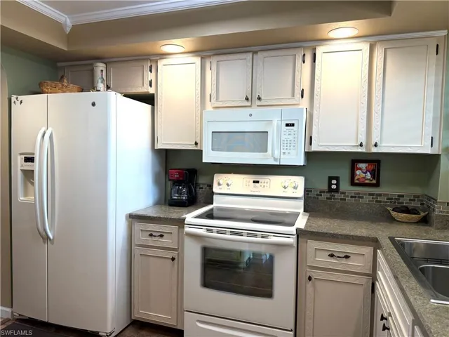 Kitchen with white appliances, dark countertops, ornamental molding, and white cabinets