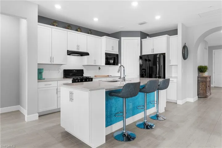 Modern kitchen featuring a gas range and oven, white shaker style cabinetry, a large center island, integrated black appliances, light-toned countertops, and wood-finish flooring. 16160 Bluestem Ln. Babcock Ranch, FL 33982
