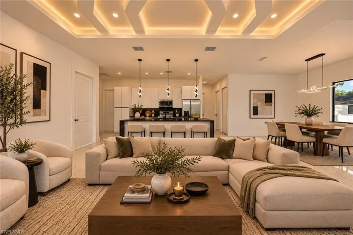 Open-concept living space featuring curved sectional seating arrangement, geometric coffered ceiling with integrated ambient lighting, natural wood-finish coffee table, and direct sightlines to a chef's kitchen with stainless steel appliances and waterfall-edge island