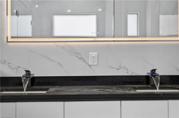 Dual vanity with black speckled countertops, under-mount sinks, and modern waterfall faucets, set against a white marble-patterned tile backsplash