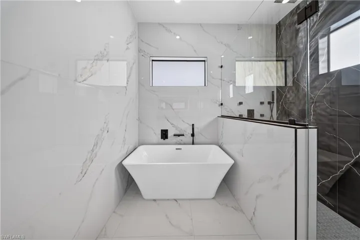 Freestanding soaking tub with wall-mounted matte black faucet, flanked by a glass-enclosed shower featuring dark stone-look tile