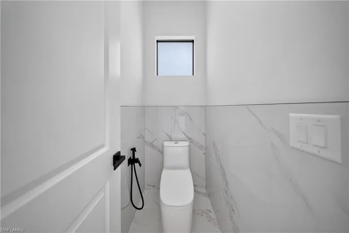 White tile flooring and wainscoting, contemporary toilet fixture, and a frosted window with black trim