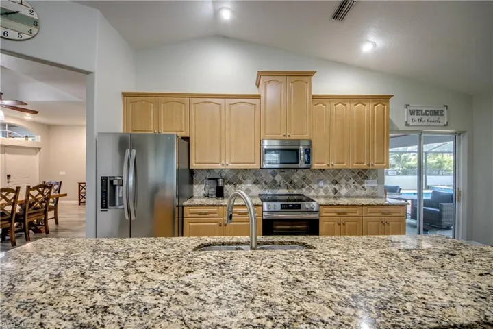 Kitchen featuring appliances with stainless steel finishes, light stone counters, vaulted ceiling, backsplash, and light brown cabinetry