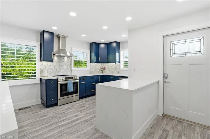 Kitchen with wall chimney exhaust hood, stainless steel electric range, a peninsula, blue cabinets, and light stone countertops - Virtually Edited Image