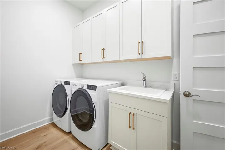 Dedicated laundry area featuring wood-finish flooring, white cabinetry with bronze hardware, a utility sink with a stone-finish countertop, and a contemporary four-panel interior door