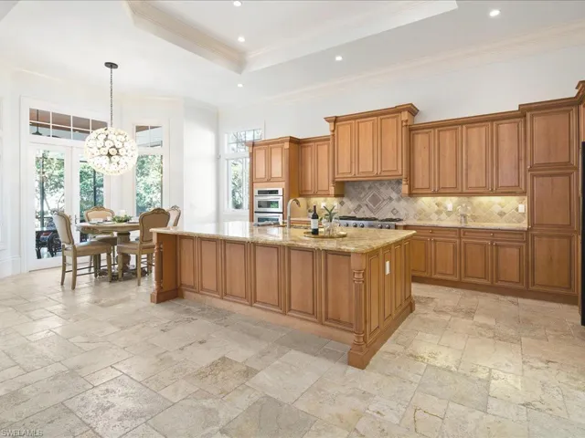 Kitchen with an inviting chandelier, decorative light fixtures, a kitchen island with sink, light stone countertops, and a raised ceiling