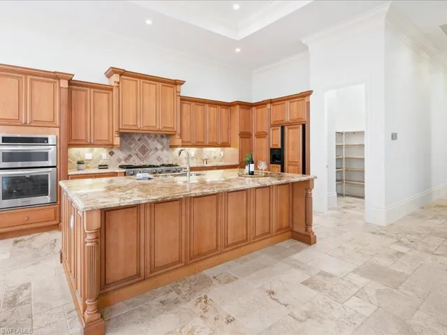 Kitchen featuring an island with sink, stainless steel double oven, light stone countertops, light tile patterned floors, and backsplash