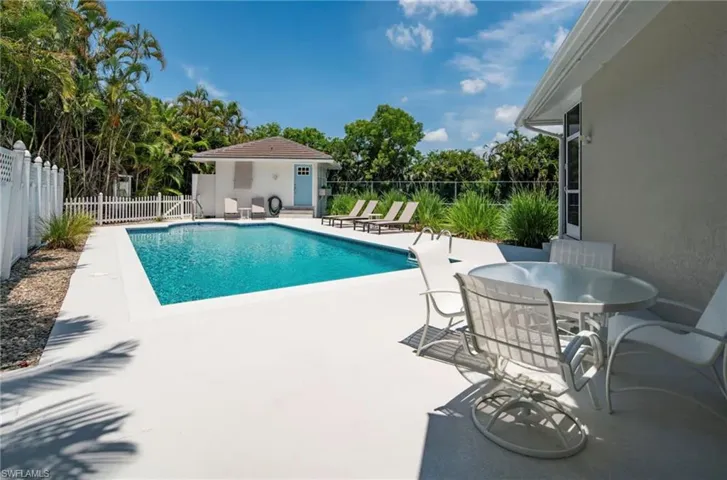 In-ground swimming pool featuring a white perimeter fence, surrounding lush tropical foliage, and a separate pool house with a light blue door