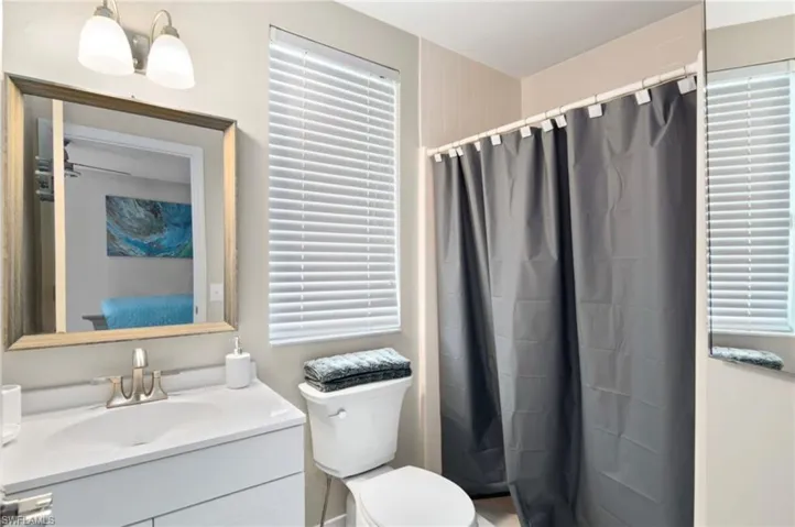 Bathroom featuring a single vanity with a rectangular sink, a framed mirror, a two-bulb vanity light fixture, a window with horizontal blinds, and a shower with a shower curtain