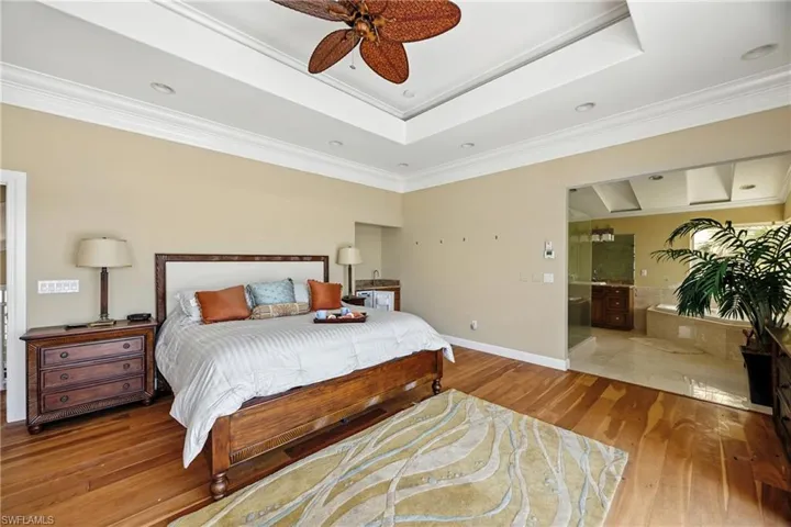 Bedroom with light wood finished floors, a tray ceiling, crown molding, ceiling fan, and ensuite bath