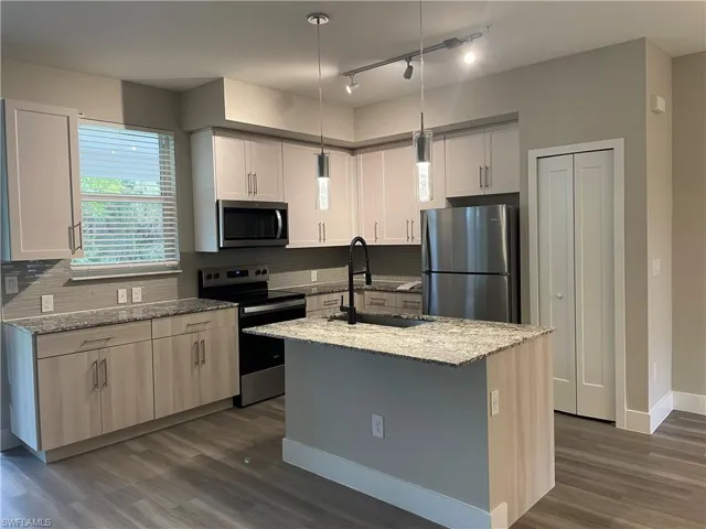 Kitchen with stainless steel appliances, light stone counters, dark wood-type flooring, a kitchen island with sink, and decorative light fixtures
