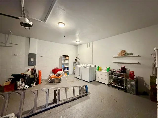 Garage with concrete block wall, electric panel, washing machine and dryer, a garage door opener, and water heater