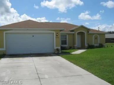 Yellow stucco exterior featuring a brown shingle roof, attached garage, concrete driveway, and a front walkway